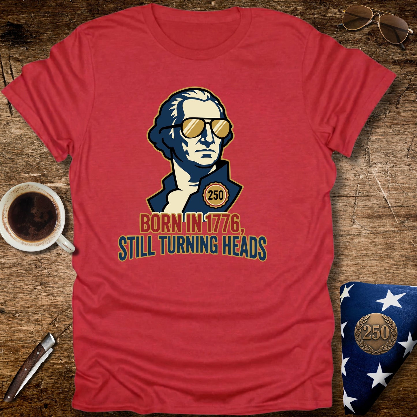 Red t-shirt with a graphic of a historical figure wearing sunglasses, text 'Born in 1776, Still Turning Heads' on a wooden surface with a cup of coffee and sunglasses.