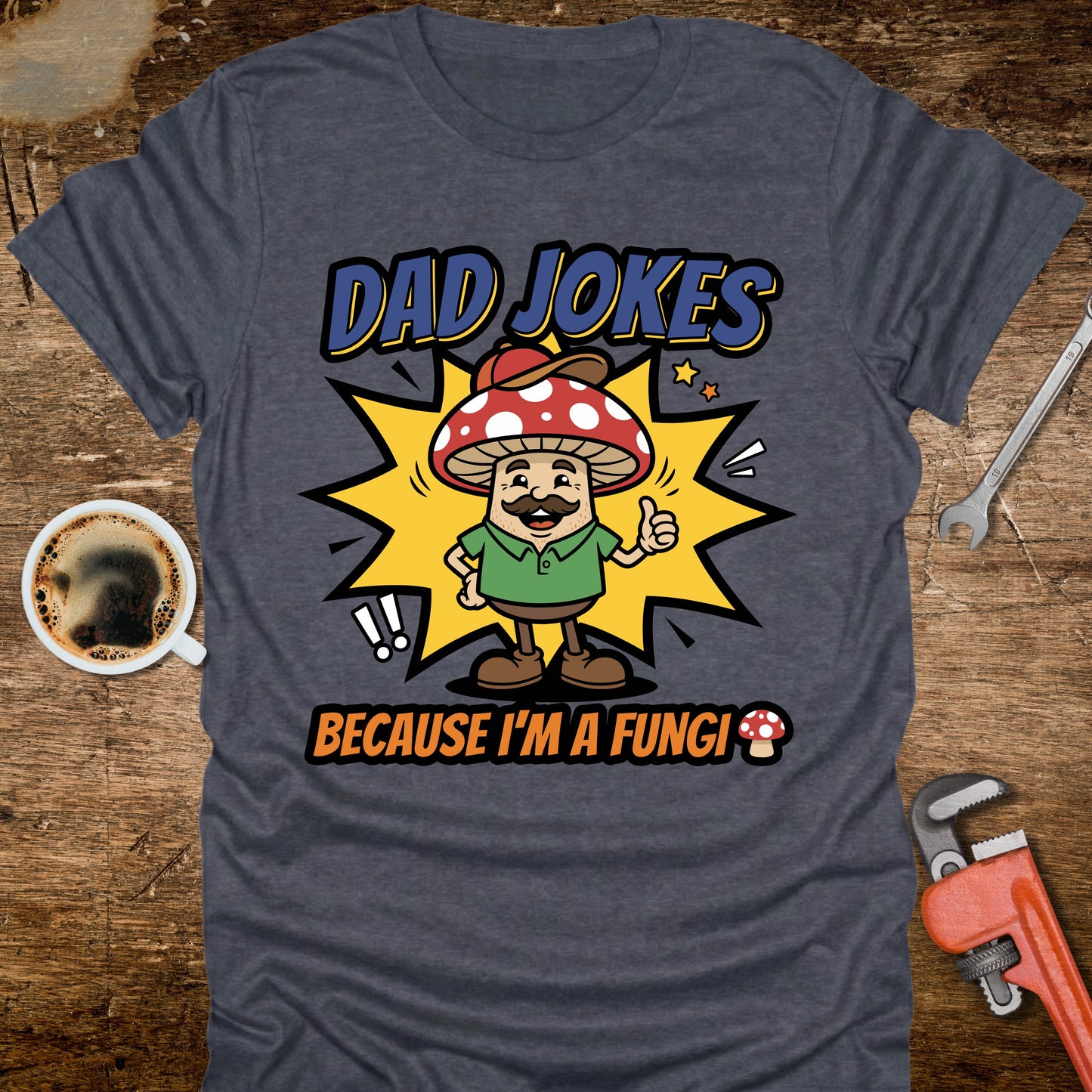 Gray t-shirt with a mushroom graphic and text 'Dad jokes because I'm a fungi' on a wooden surface with coffee and tools.