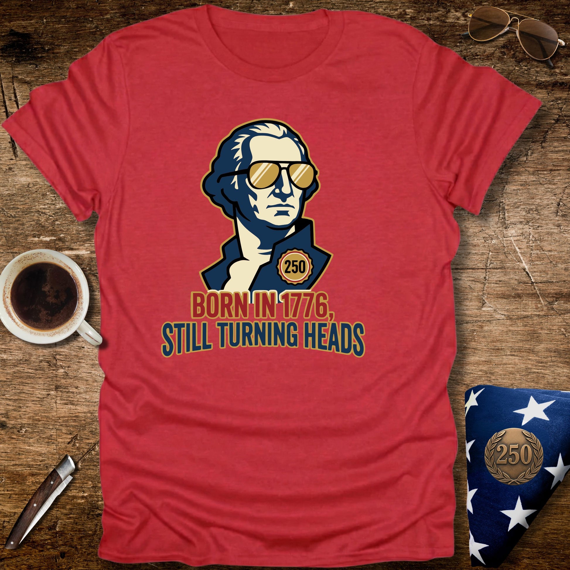 Red t-shirt with a graphic of a historical figure wearing sunglasses, text 'Born in 1776, Still Turning Heads' on a wooden surface with a cup of coffee and sunglasses.