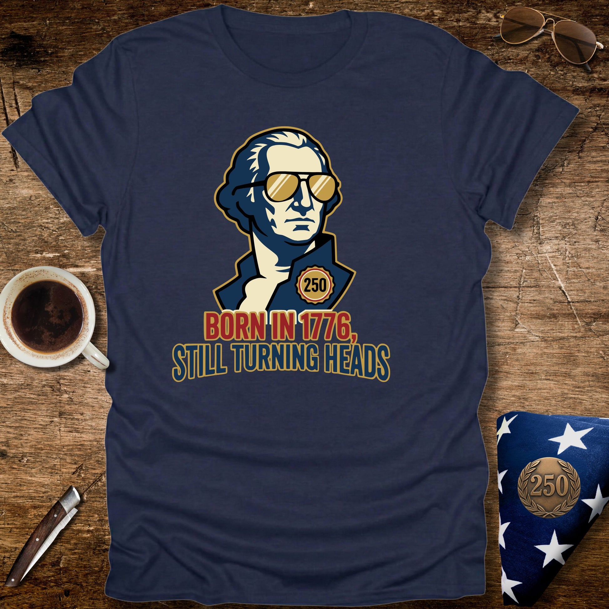 Navy blue t-shirt with a graphic of a historical figure wearing sunglasses and text 'Born in 1776, Still Turning Heads' on a wooden surface.