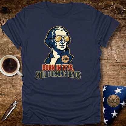 Navy blue t-shirt with a graphic of a historical figure wearing sunglasses and text 'Born in 1776, Still Turning Heads' on a wooden surface.