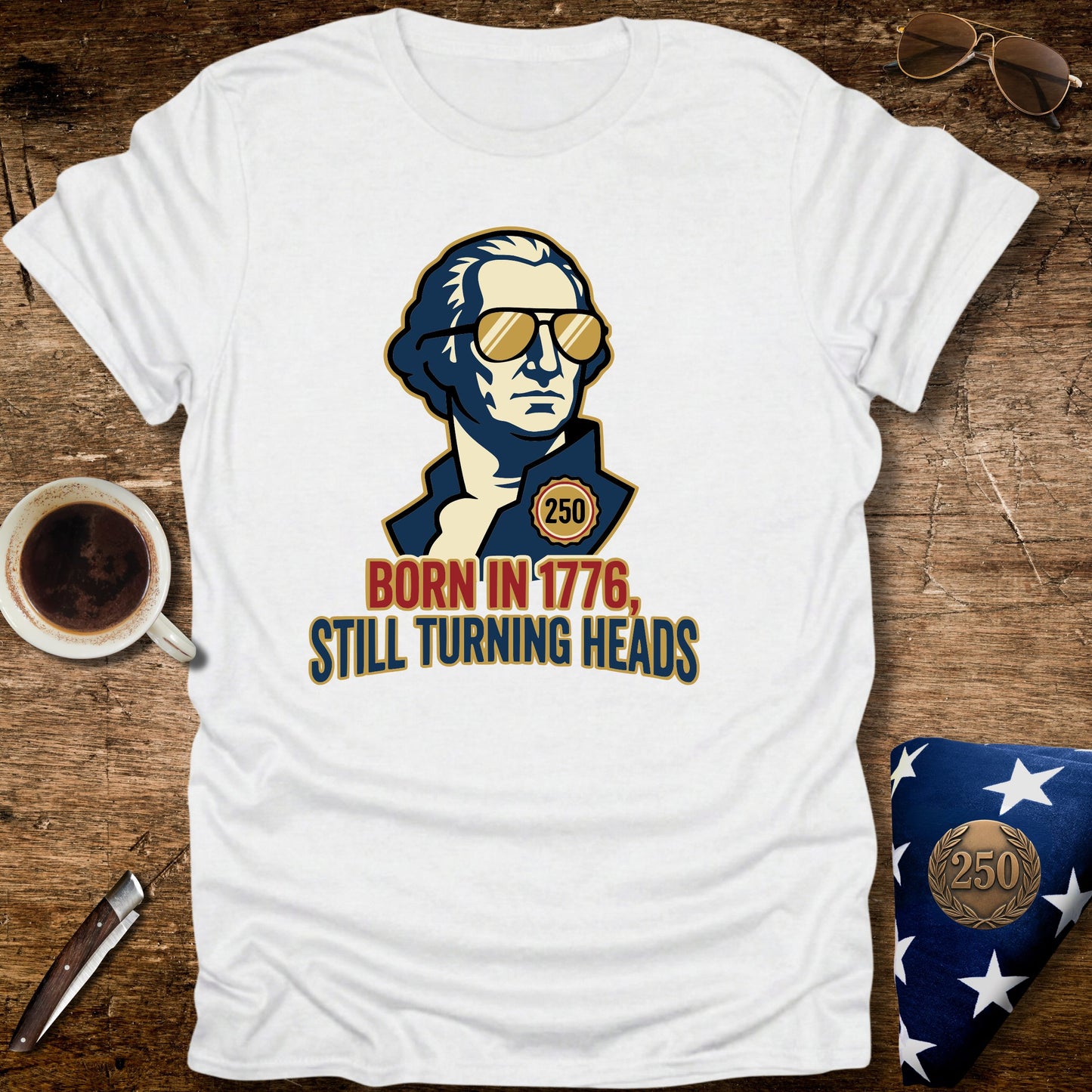 White t-shirt with graphic design of a person wearing sunglasses and text 'Born in 1776, Still Turning Heads' on a wooden surface.