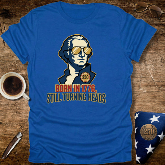 Blue t-shirt with a graphic design of a person wearing sunglasses, text 'Born in 1776, Still Turning Heads' on a wooden surface with a cup and flag.