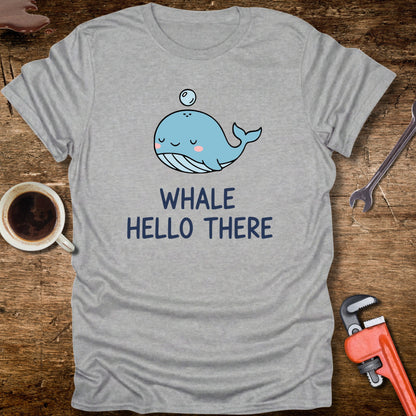 Gray t-shirt with a cartoon whale and 'Whale Hello There' text on a wooden surface with a coffee cup and tools.