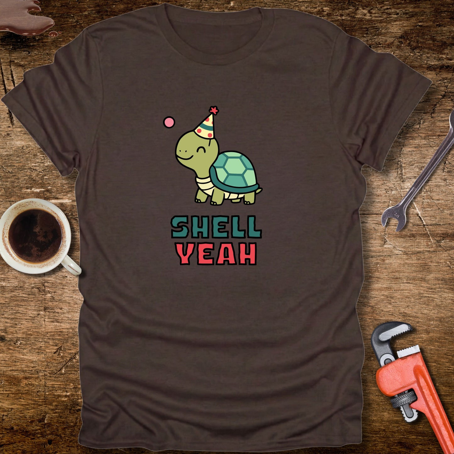 Brown t-shirt with a turtle graphic and 'Shell Yeah' text on a wooden surface with tools and a cup.