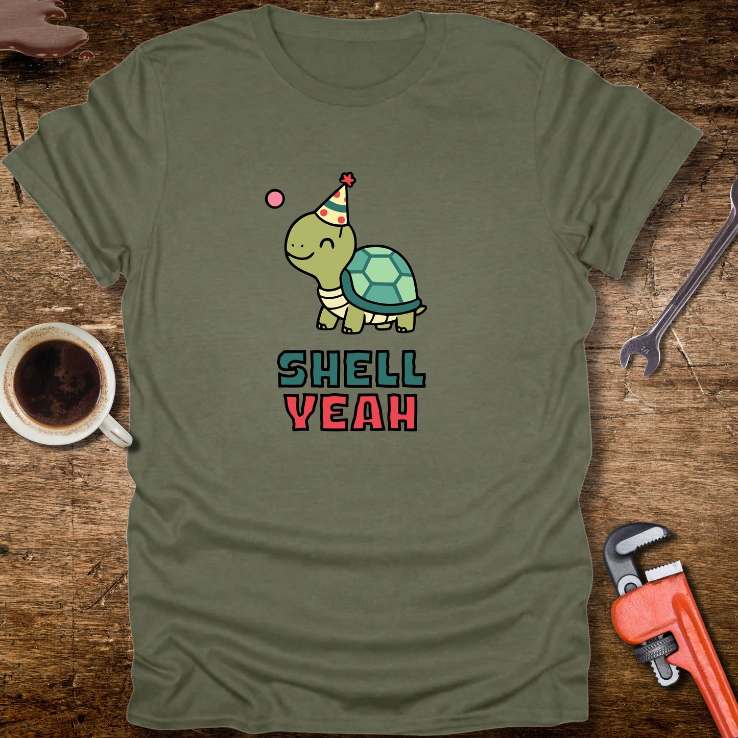 Green t-shirt with a turtle graphic and 'Shell Yeah' text on a wooden surface with tools and a cup.