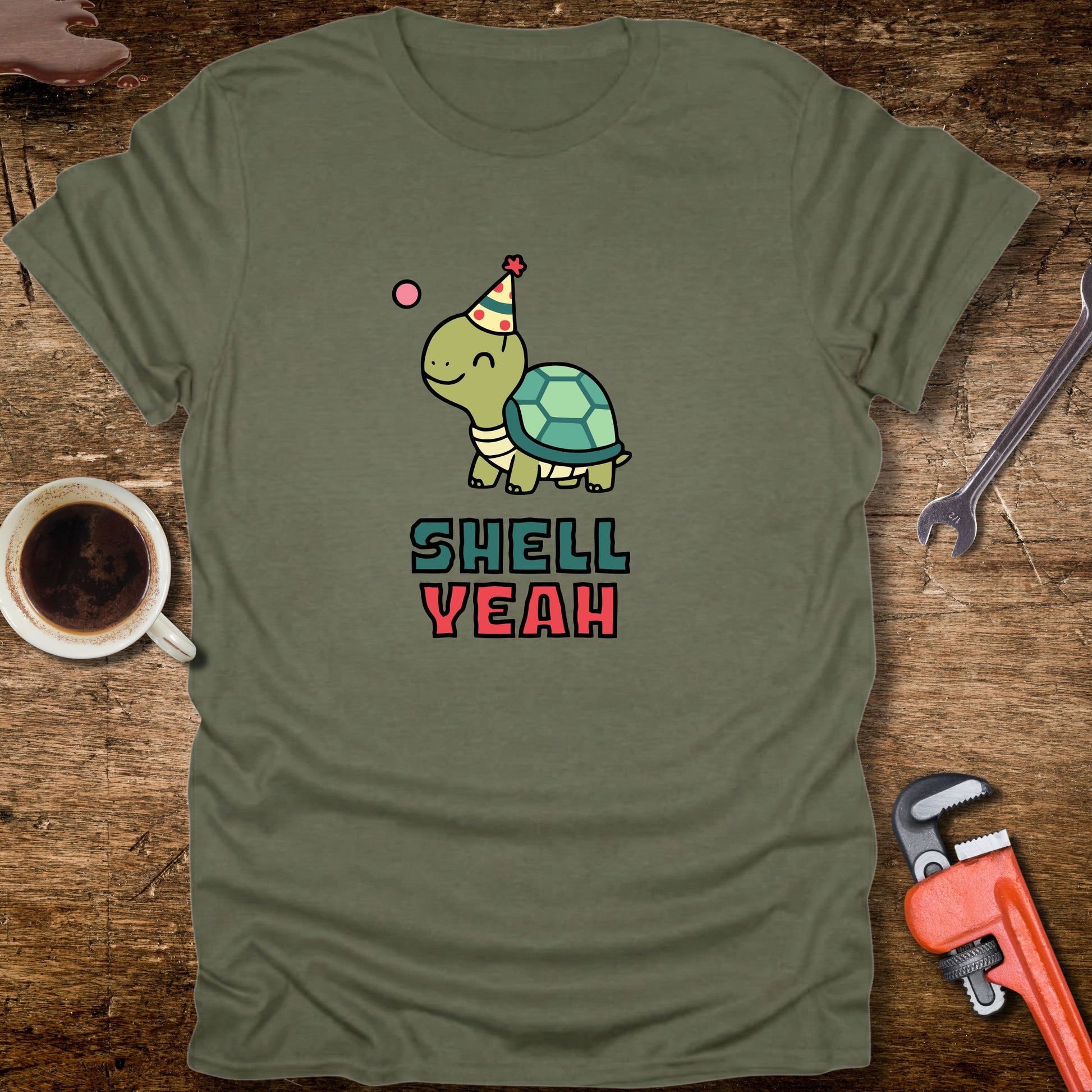 Green t-shirt with a turtle graphic and 'Shell Yeah' text on a wooden surface with tools and a cup.