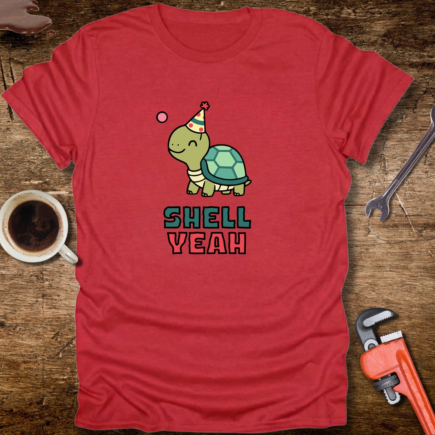 Red t-shirt with a turtle graphic and 'Shell Yeah' text on a wooden surface with tools and a cup.