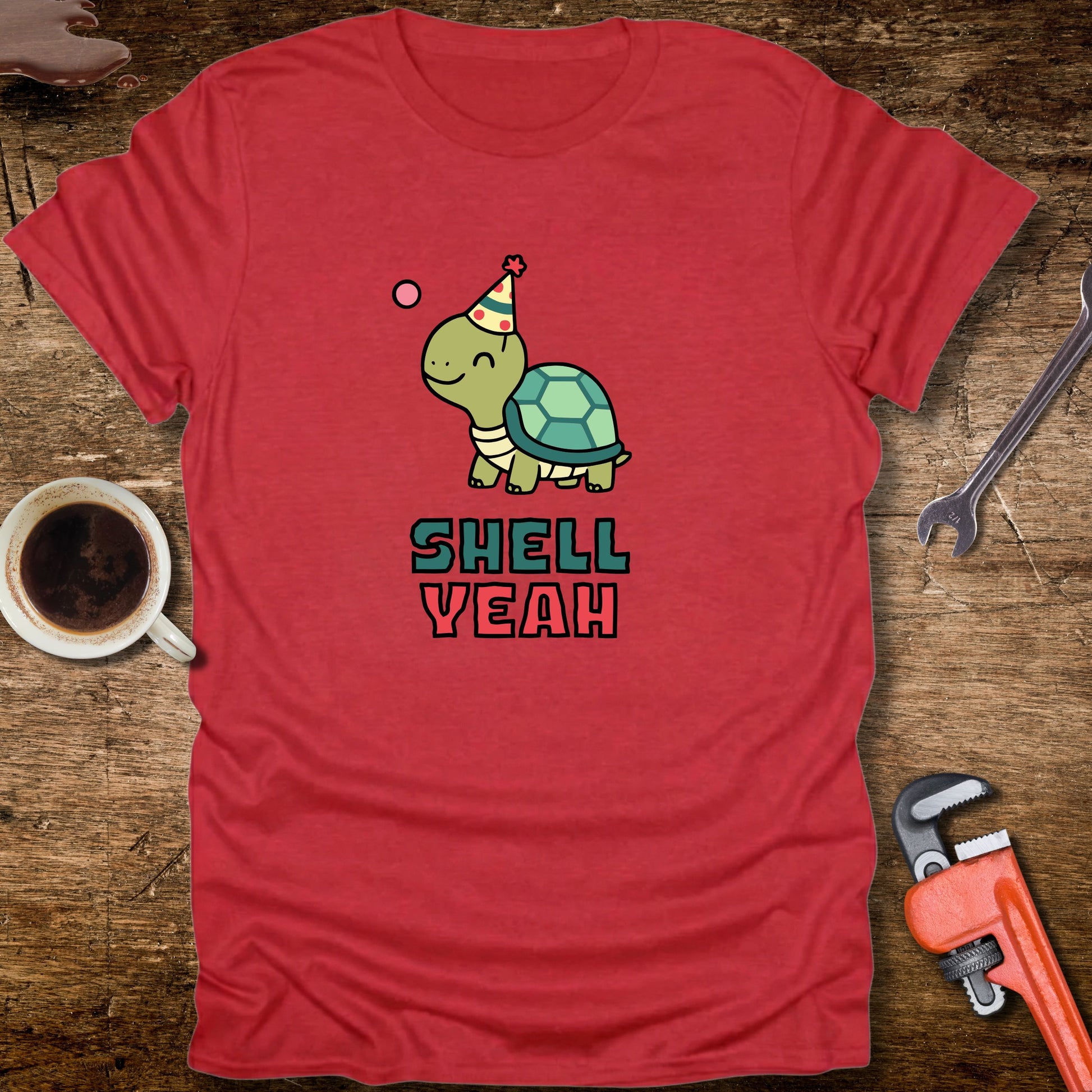 Red t-shirt with a turtle graphic and 'Shell Yeah' text on a wooden surface with tools and a cup.