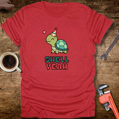 Red t-shirt with a turtle graphic and 'Shell Yeah' text on a wooden surface with tools and a cup.