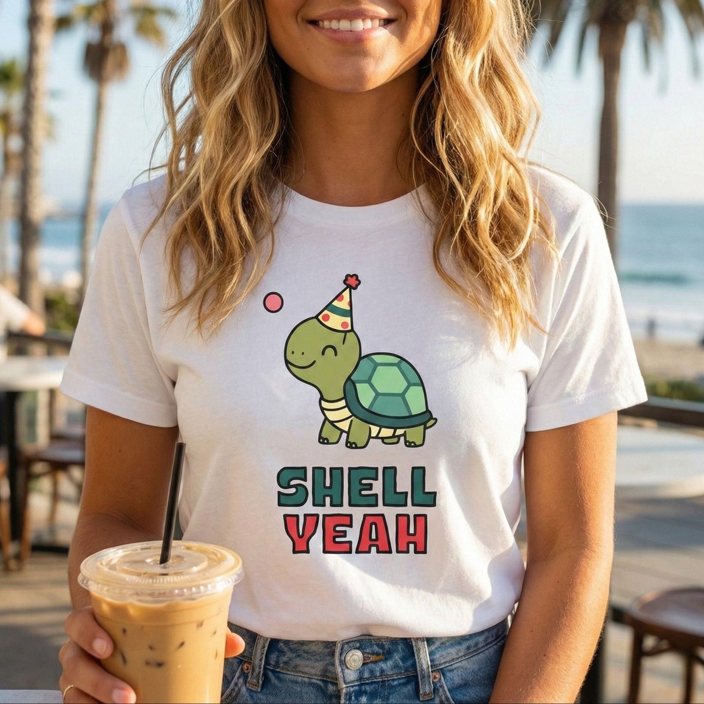 Woman wearing a white t-shirt with a turtle graphic and 'Shell Yeah' text, holding a drink by the beach.