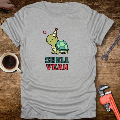 Gray t-shirt with a turtle graphic and 'Shell Yeah' text on a wooden surface with tools and a cup.