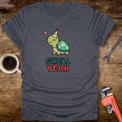 Gray t-shirt with a turtle graphic and 'Shell Yeah' text on a wooden surface with tools and a cup.