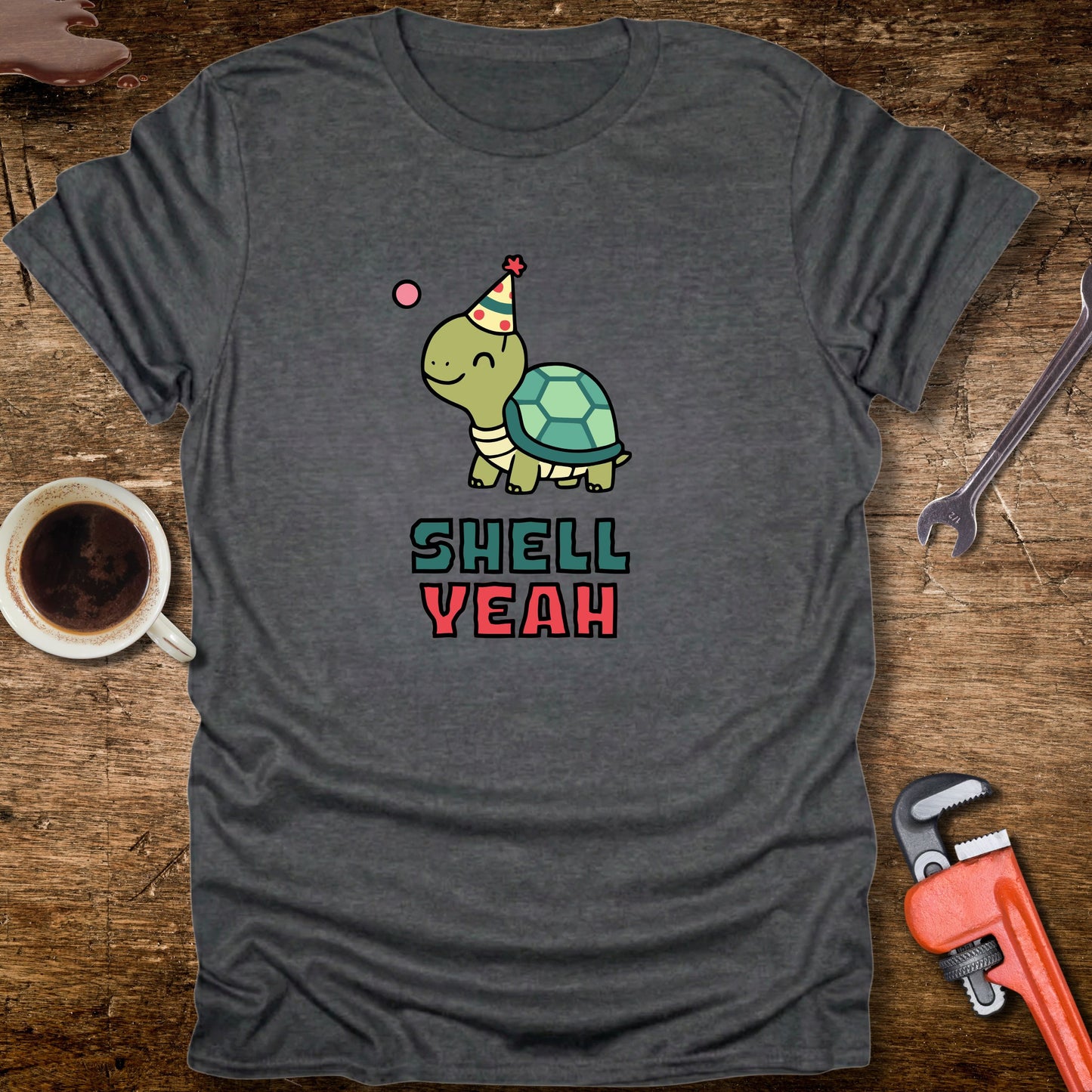 Gray t-shirt with a turtle graphic and 'Shell Yeah' text on a wooden surface with tools and a cup.
