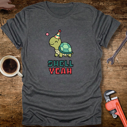 Gray t-shirt with a turtle graphic and 'Shell Yeah' text on a wooden surface with tools and a cup.