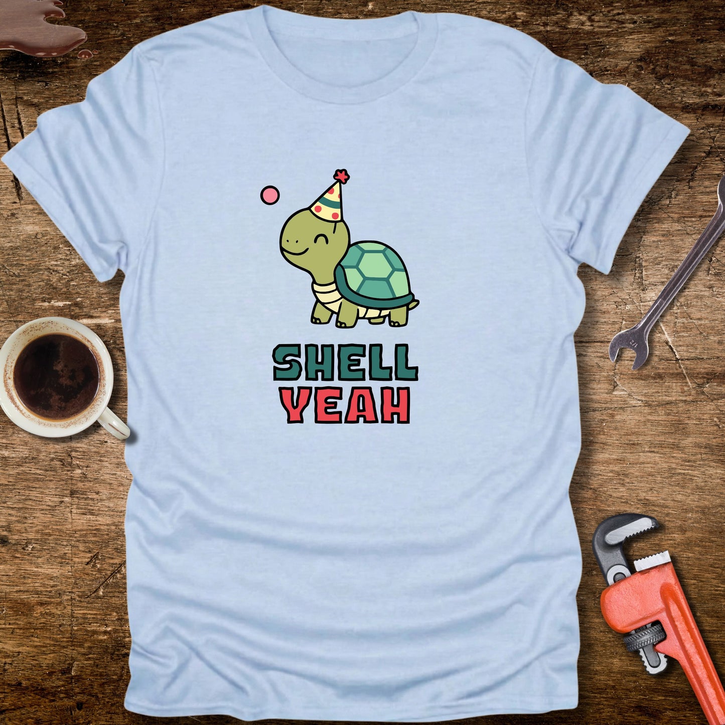 Light blue t-shirt with a turtle graphic and 'Shell Yeah' text on a wooden surface with tools and a cup.