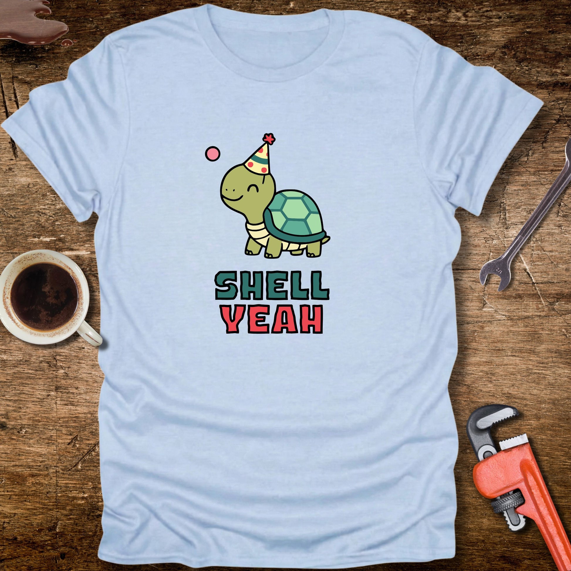 Light blue t-shirt with a turtle graphic and 'Shell Yeah' text on a wooden surface with tools and a cup.