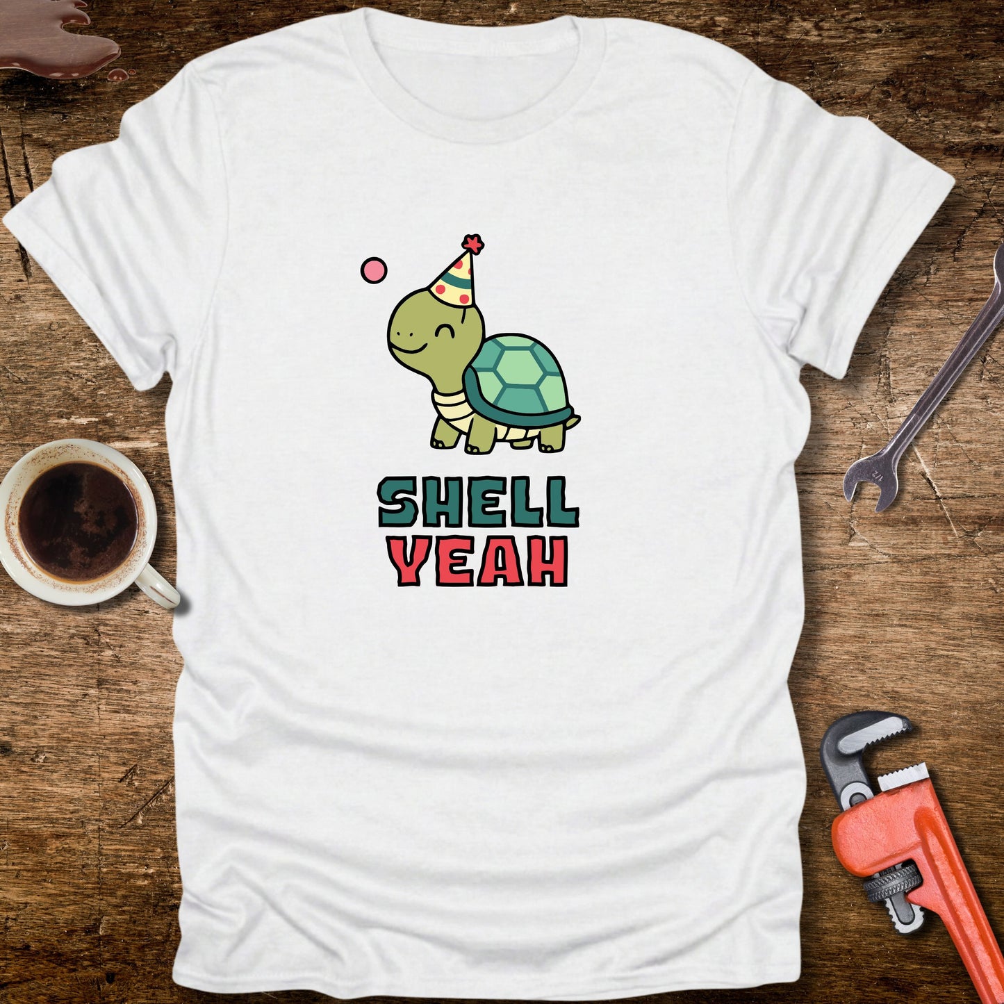 White t-shirt with a turtle graphic and 'Shell Yeah' text on a wooden surface with tools and a cup.