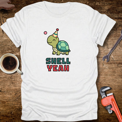 White t-shirt with a turtle graphic and 'Shell Yeah' text on a wooden surface with tools and a cup.