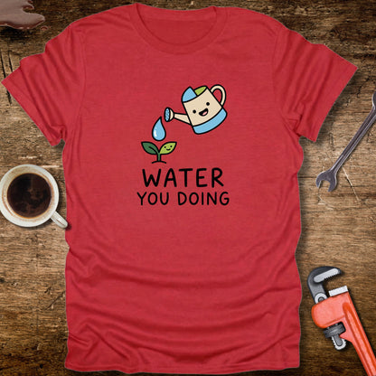Red t-shirt with a cartoon watering can and 'Water You Doing' text on a wooden surface with tools and a cup.