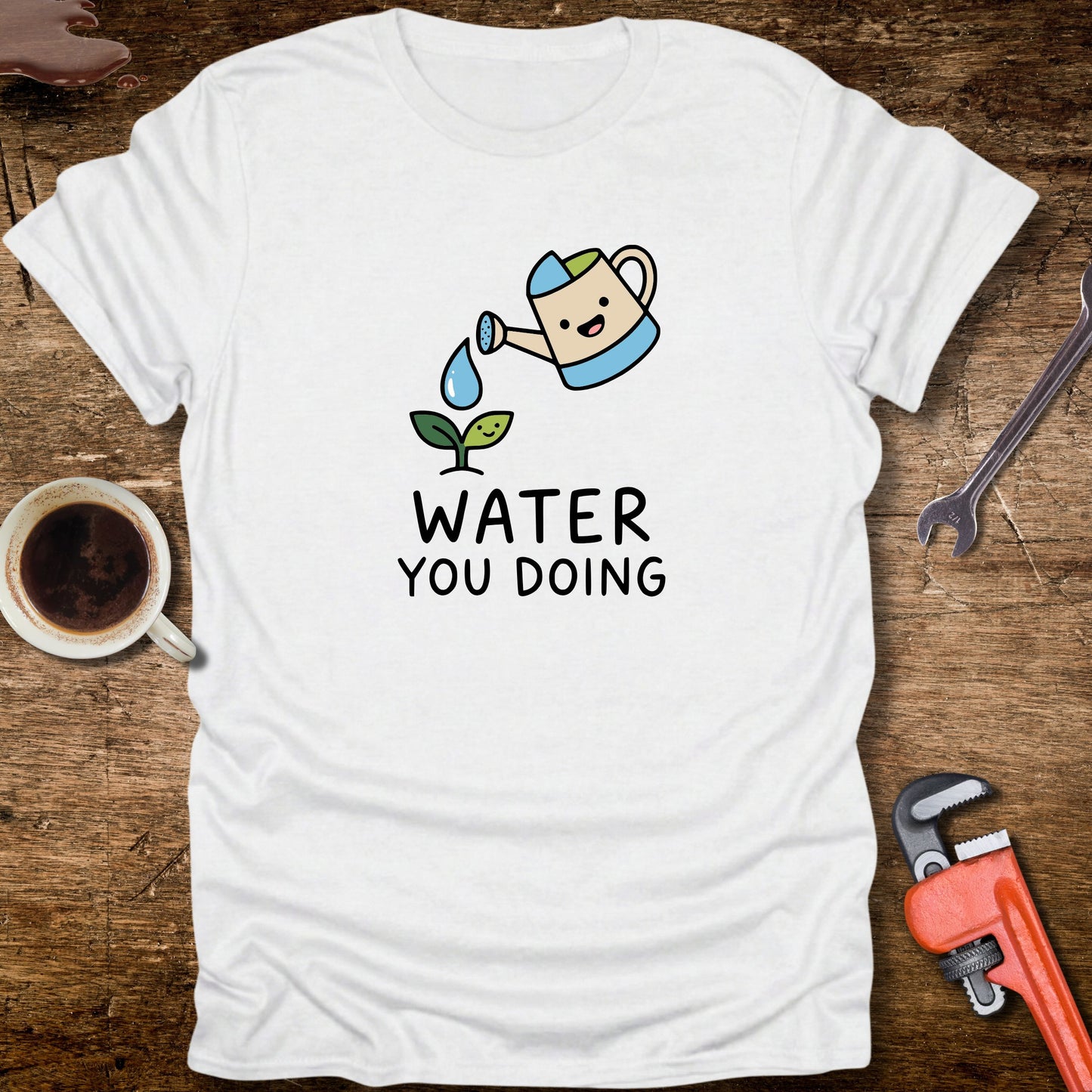 White t-shirt with a cartoon watering can and 'Water You Doing' text on a wooden surface with tools and a cup.