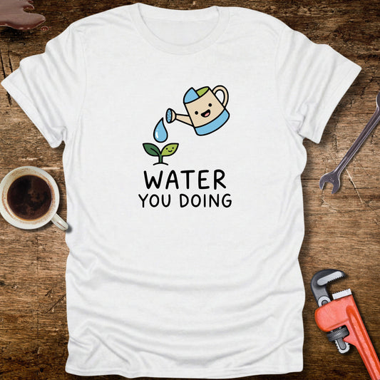 White t-shirt with a cartoon watering can and 'Water You Doing' text on a wooden surface with tools and a cup.