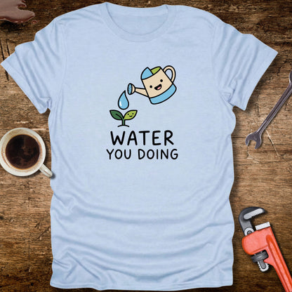 Light blue t-shirt with cartoon watering can and plant design on a wooden surface with tools and a cup.