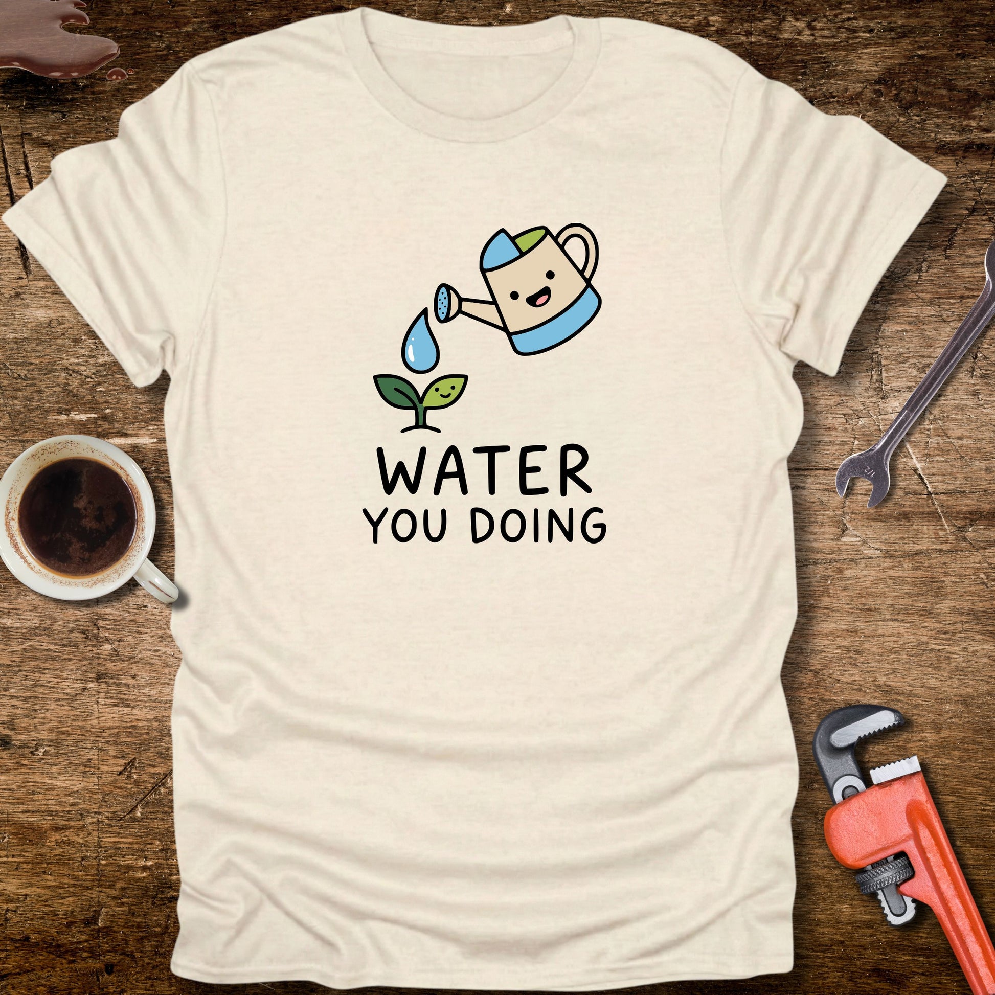 Beige t-shirt with a graphic design and text on a wooden surface with tools and a cup.