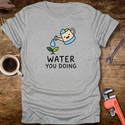 Gray t-shirt with a cartoon watering can and 'Water You Doing' text on a wooden surface with tools and a cup.
