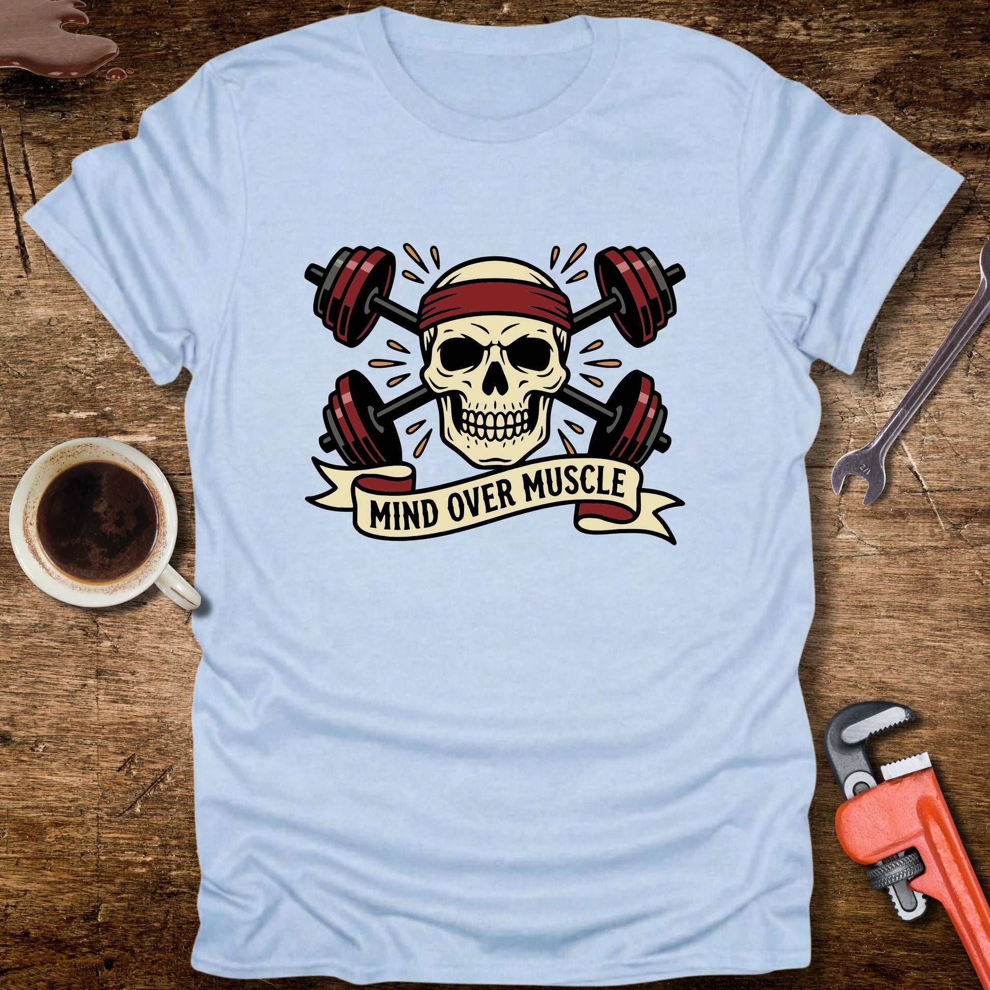 Light blue t-shirt with skull and barbell design on a wooden surface with coffee and tools.