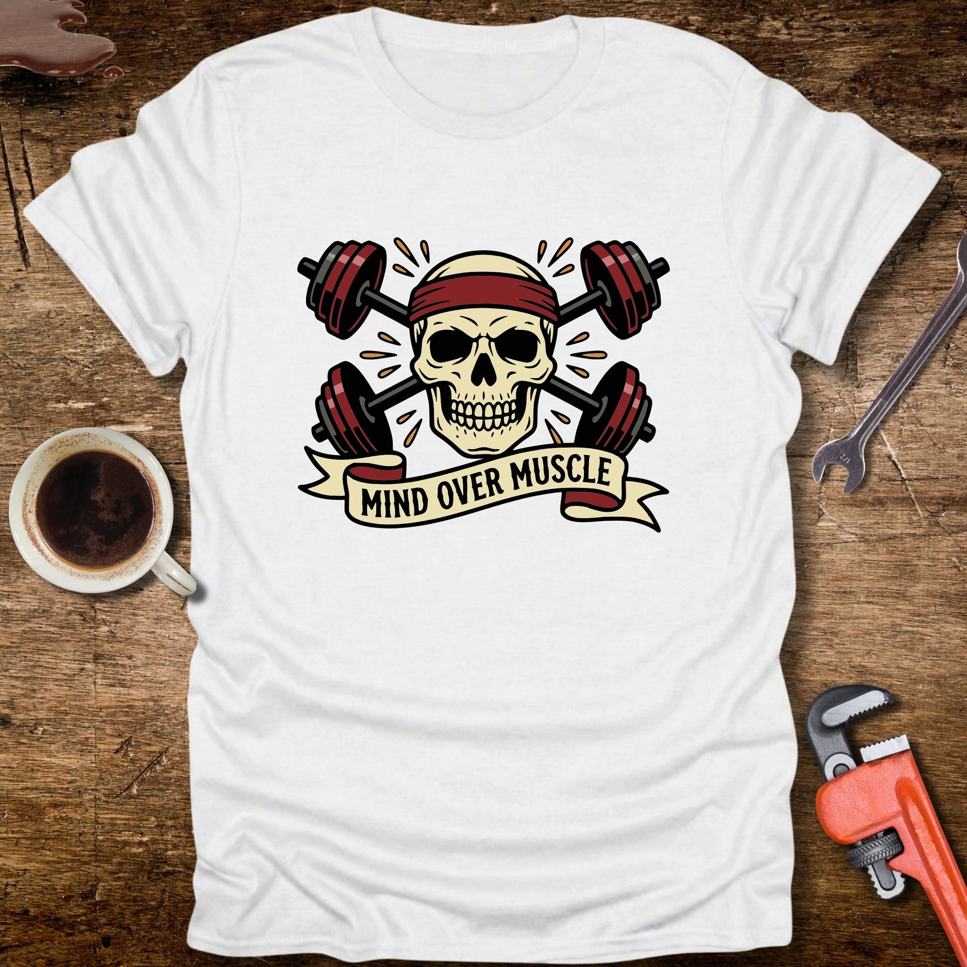 White t-shirt with skull and barbell design on a wooden surface with coffee and tools.