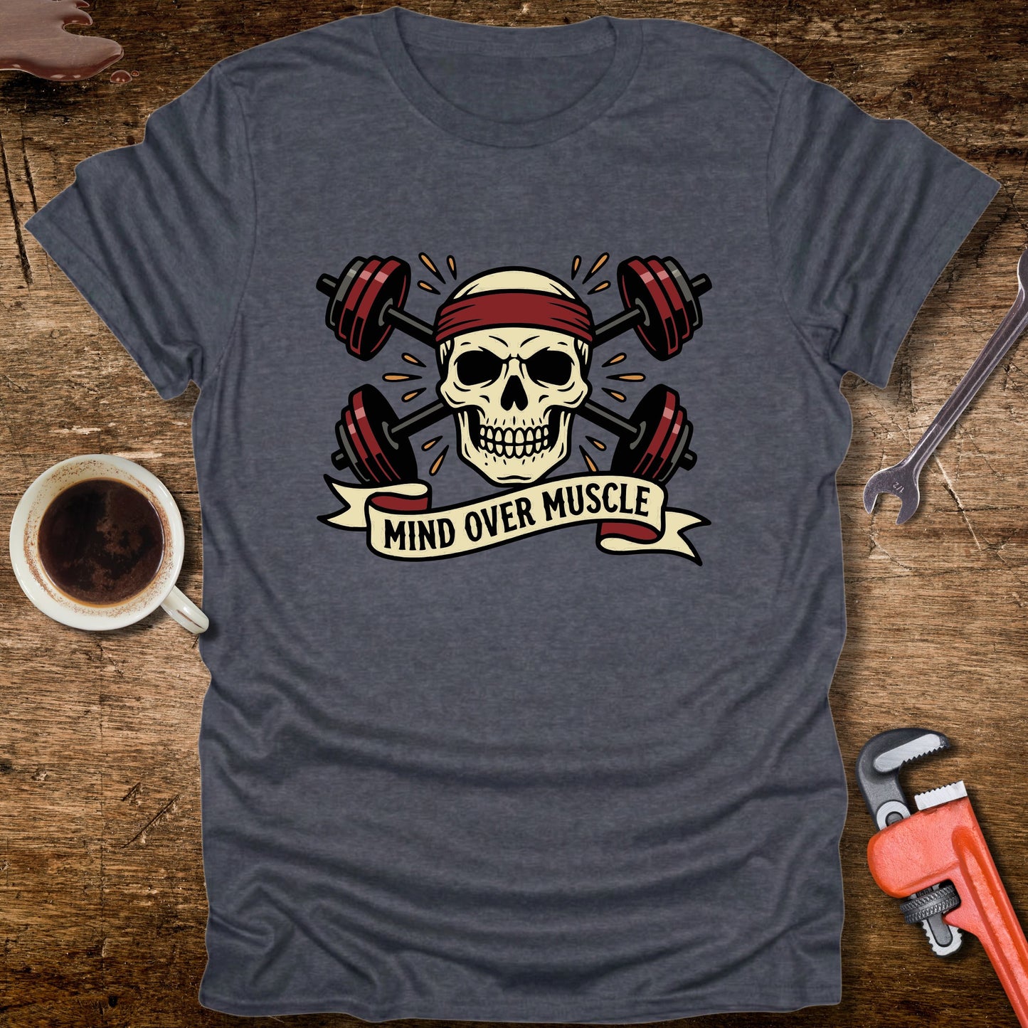 Gray t-shirt with skull and barbell design on a wooden surface with coffee and tools.