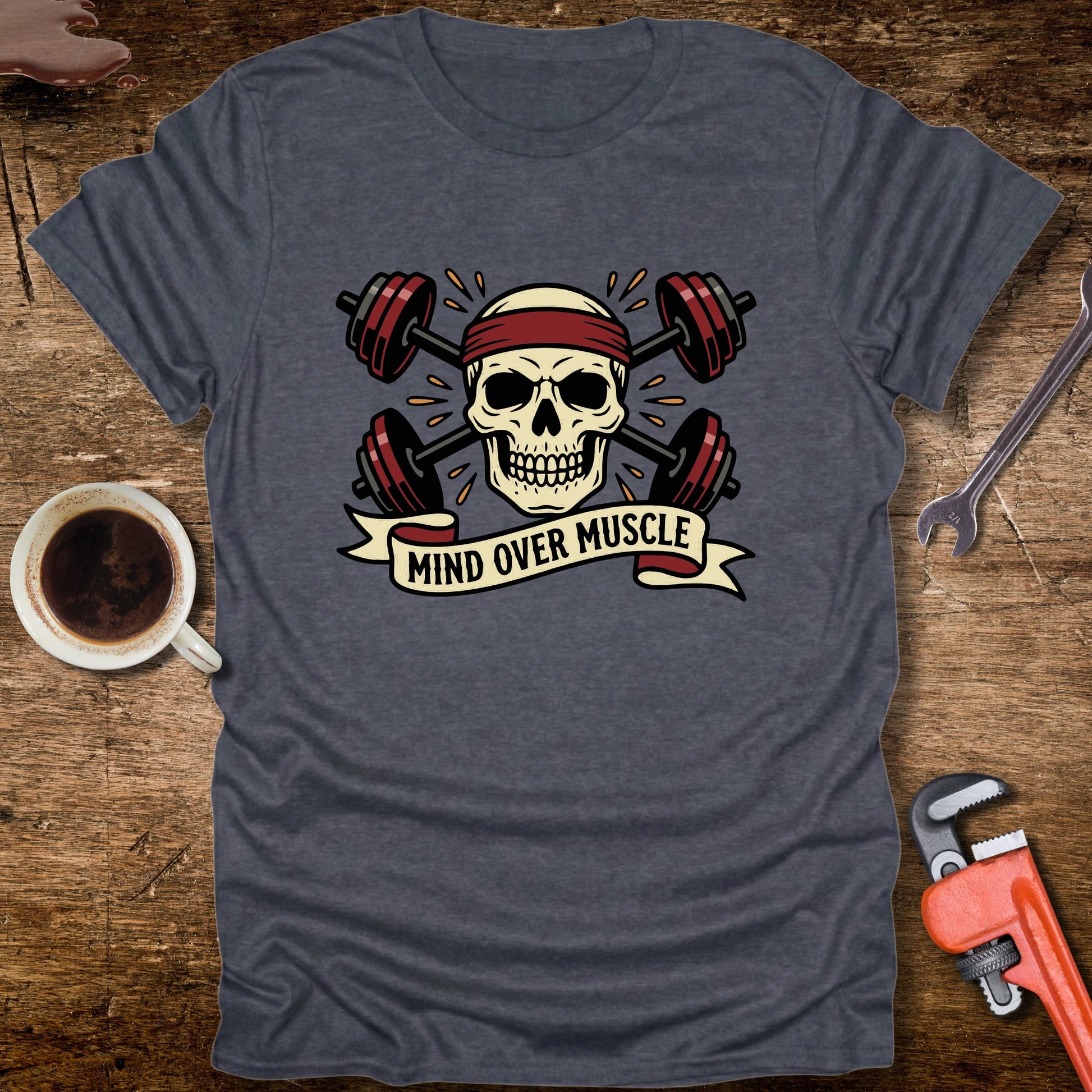 Gray t-shirt with skull and barbell design on a wooden surface with coffee and tools.