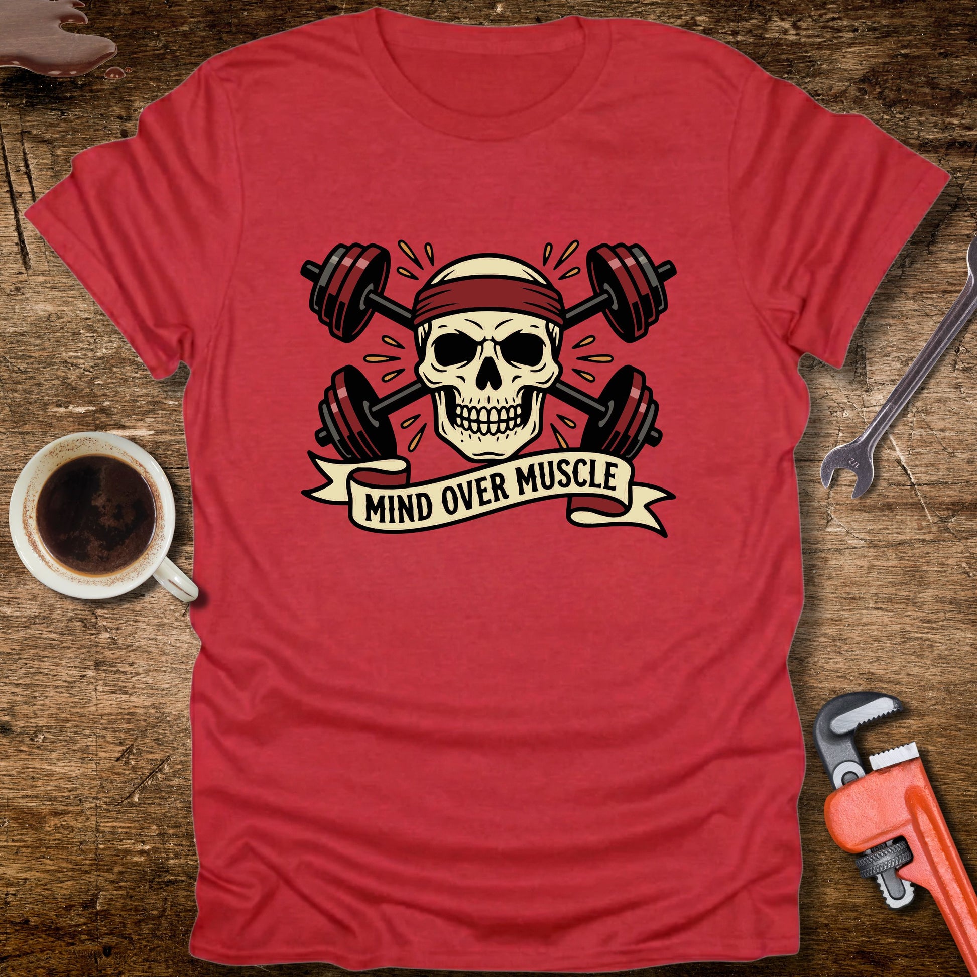 Red t-shirt with skull and barbell graphic and 'Mind Over Muscle' text on a wooden surface with tools and a cup.