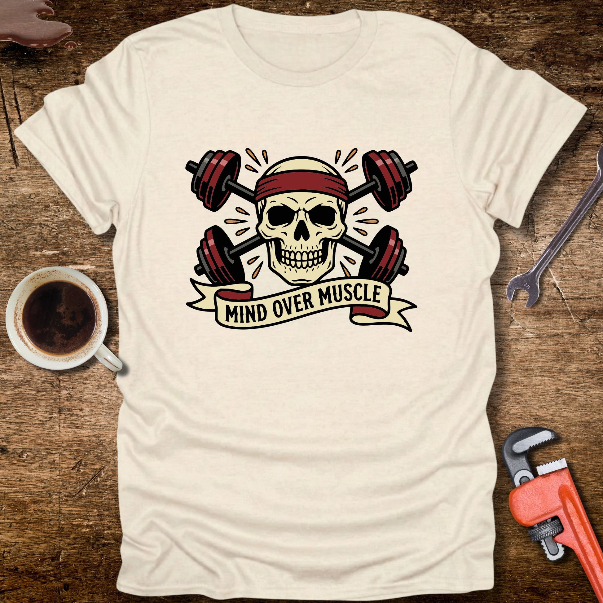 White t-shirt with skull and barbell graphic and 'Mind Over Muscle' text on a wooden surface with coffee and tools.