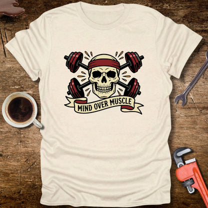 White t-shirt with skull and barbell graphic and 'Mind Over Muscle' text on a wooden surface with coffee and tools.