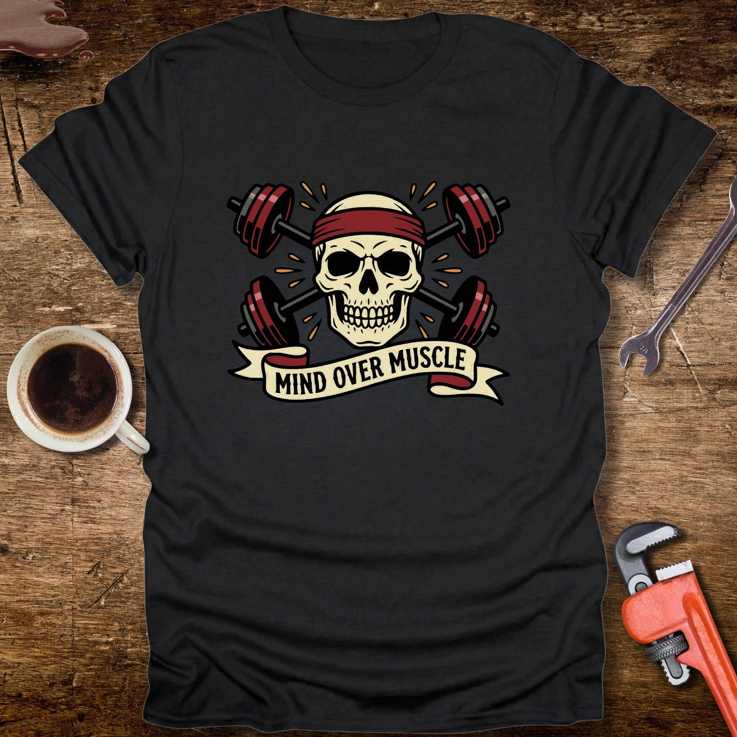Black t-shirt with skull and barbell design on a wooden surface with coffee and tools.