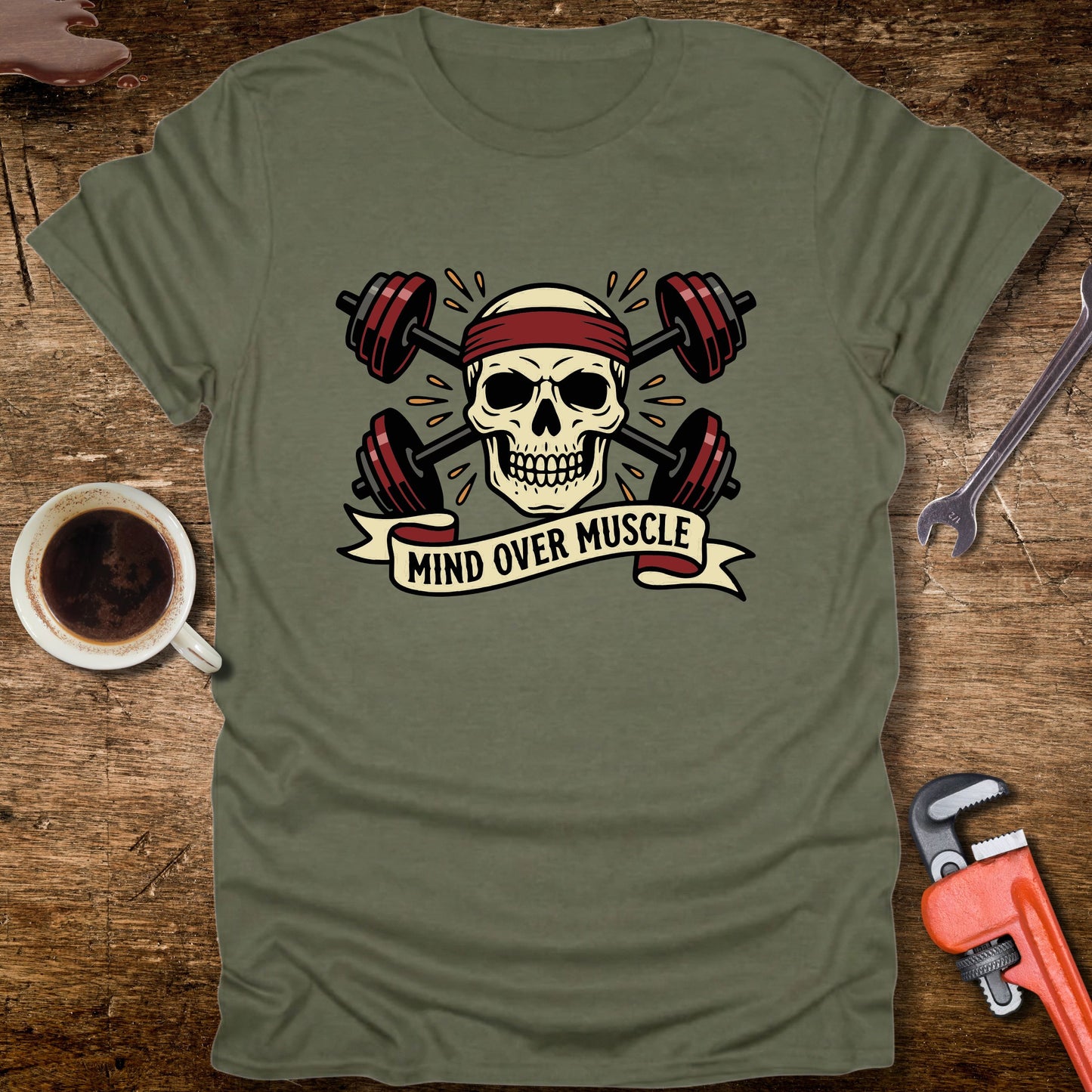 Green t-shirt with skull and barbell design on a wooden surface with coffee and tools.