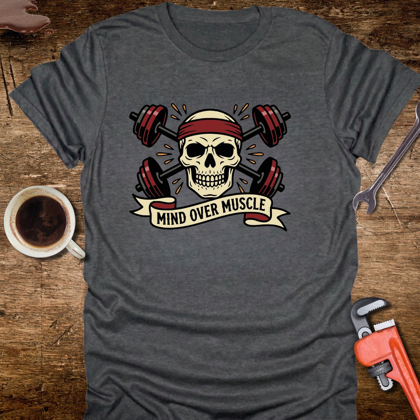 Gray t-shirt with skull and barbell design on a wooden surface with coffee and tools.