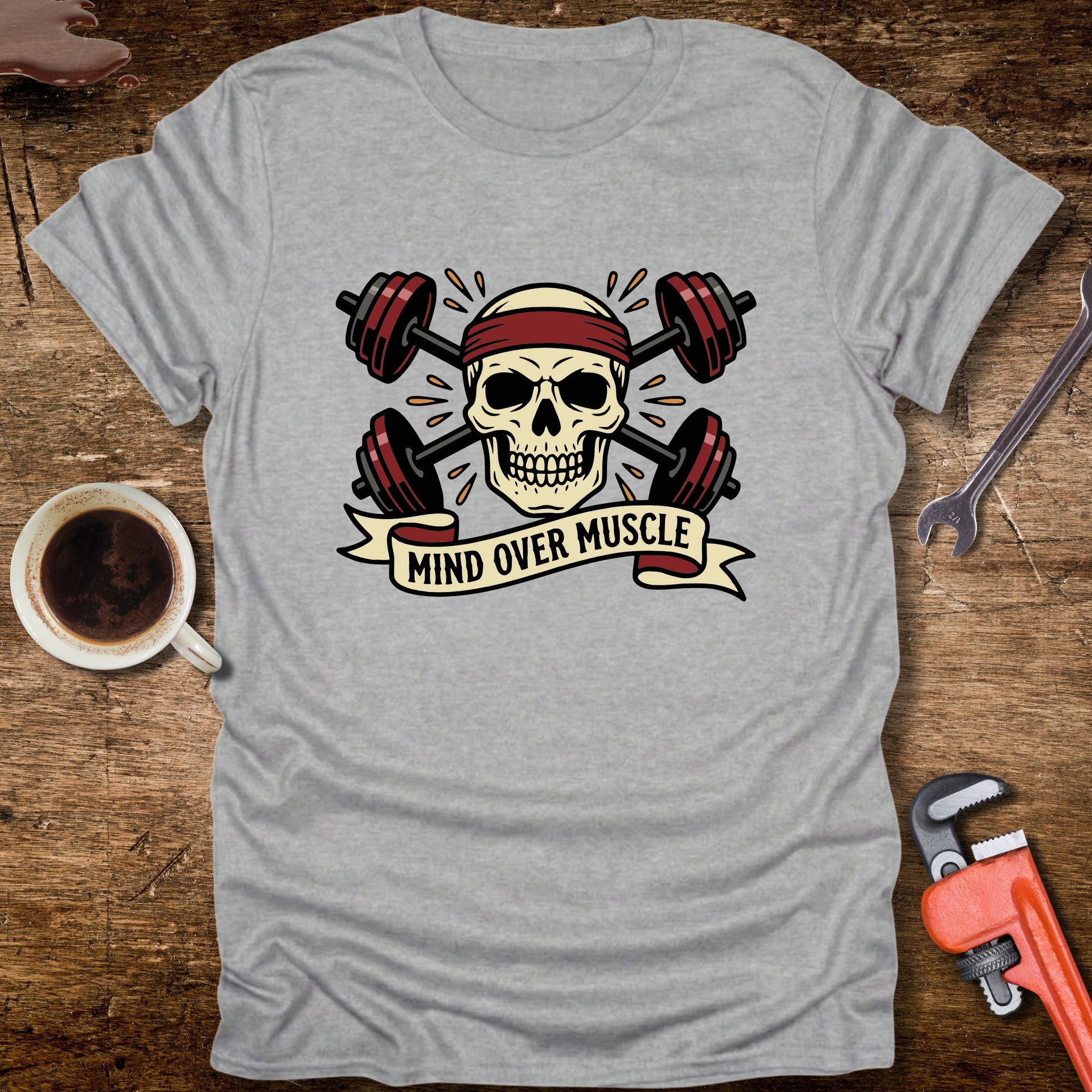 Gray t-shirt with skull and barbell design on a wooden surface with coffee and tools.