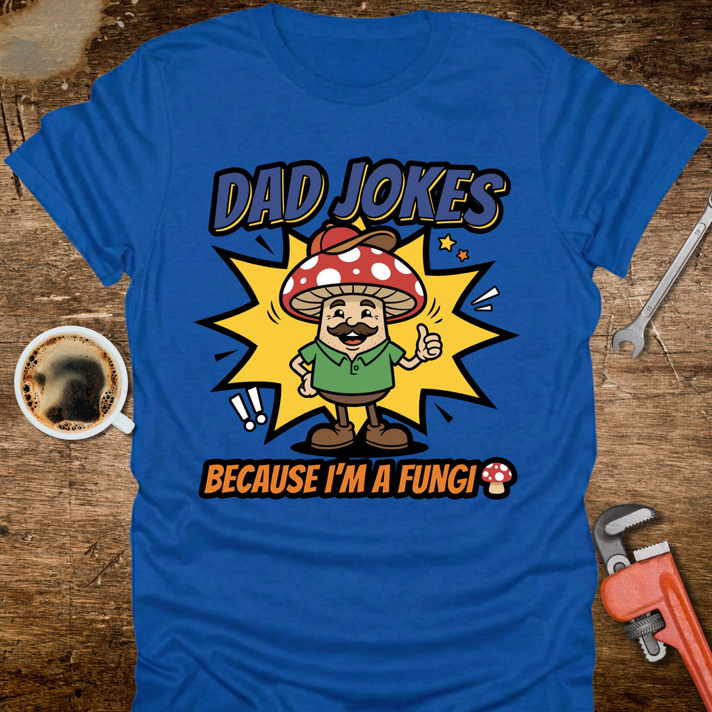 Blue t-shirt with a cartoon mushroom character and text on a wooden surface with tools and a coffee cup.