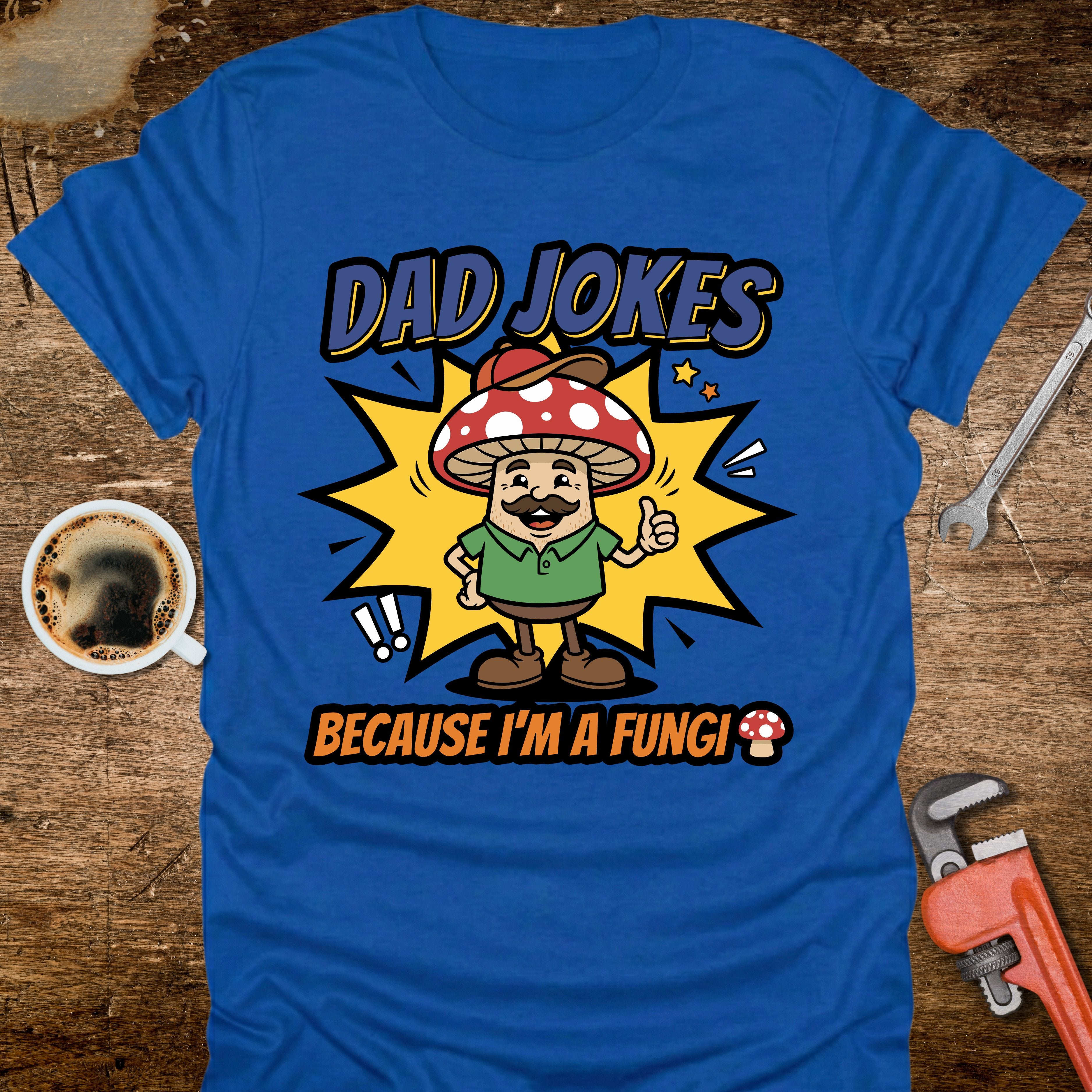 Blue t-shirt with a cartoon mushroom character and text on a wooden surface with tools and a coffee cup.