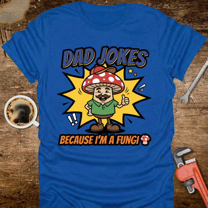 Blue t-shirt with a cartoon mushroom character and text on a wooden surface with tools and a coffee cup.