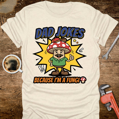 White t-shirt with 'Dad Jokes Because I'm a Fungi' graphic on a wooden surface with coffee and tools.