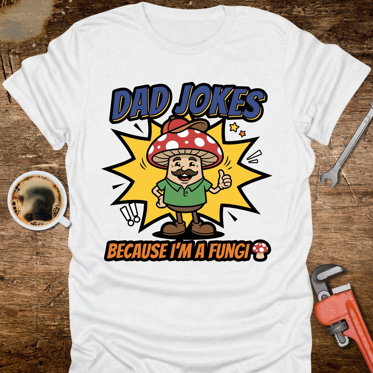 White t-shirt with 'Dad Jokes Because I'm a Fungi' graphic on a wooden surface with coffee and tools.