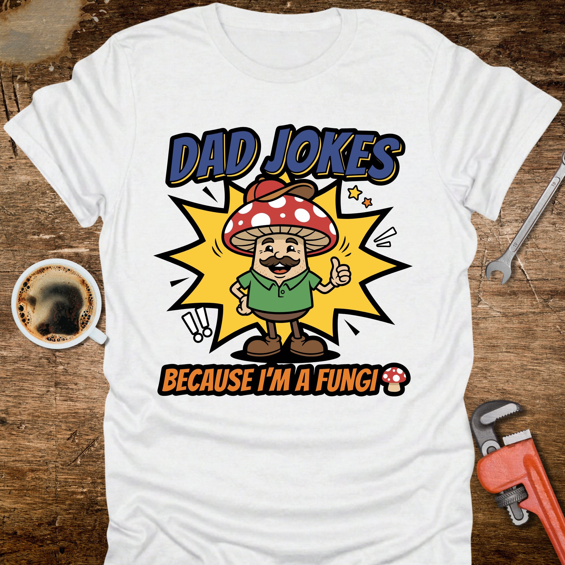 White t-shirt with 'Dad Jokes Because I'm a Fungi' graphic on a wooden surface with coffee and tools.