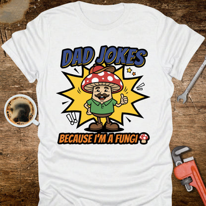 White t-shirt with 'Dad Jokes Because I'm a Fungi' graphic on a wooden surface with coffee and tools.