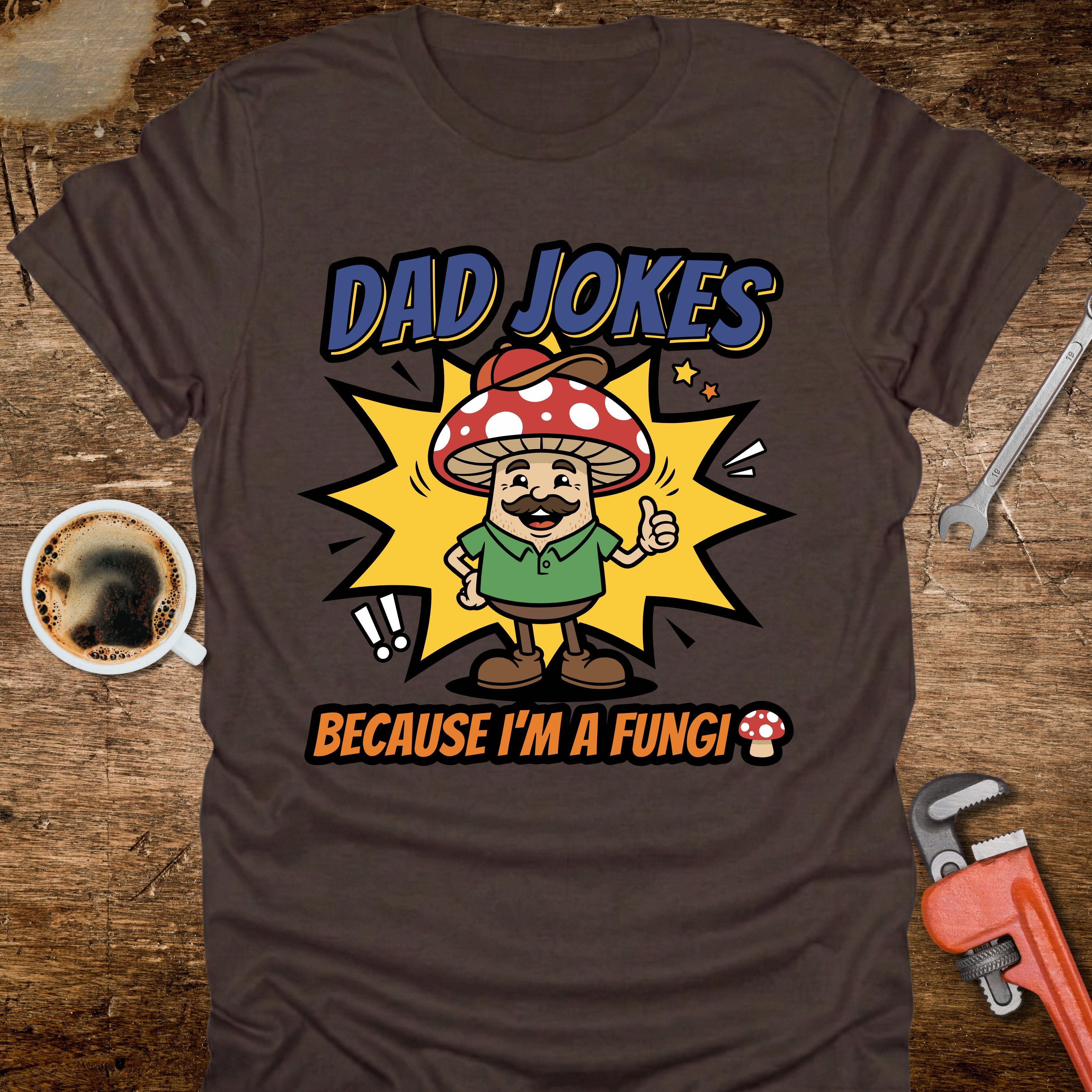 Brown t-shirt with a cartoon mushroom character and text on a wooden surface with coffee and tools.