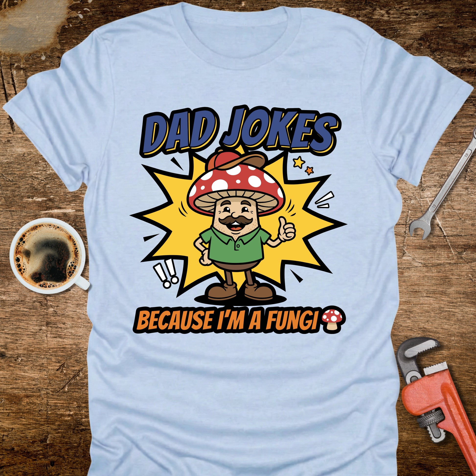 Light blue t-shirt with a cartoon mushroom character and text on a wooden surface with a coffee cup and wrench.