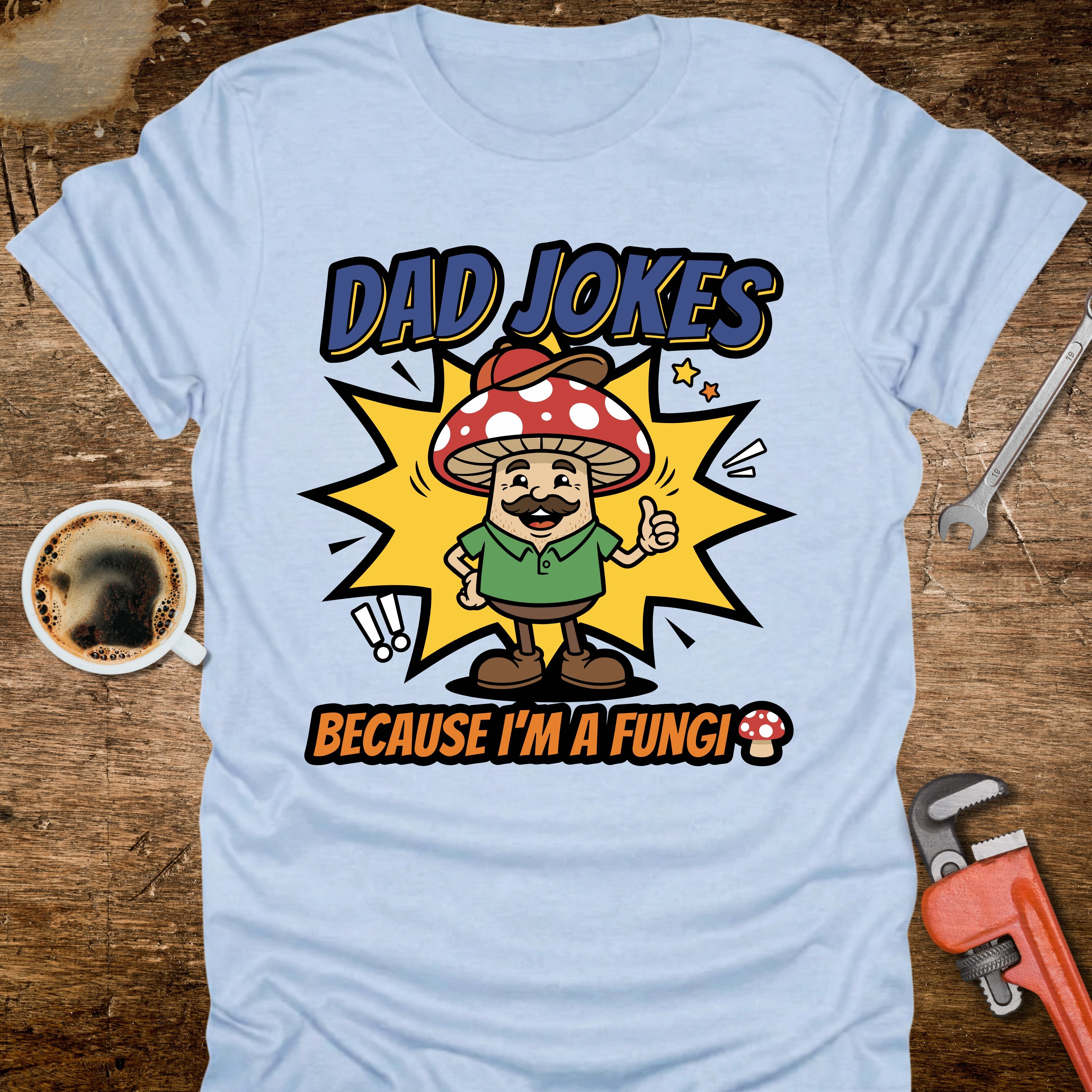 Light blue t-shirt with a cartoon mushroom character and text on a wooden surface with a coffee cup and wrench.